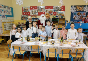 Grupa dzieci ubranych odświętnie, znajdujących się w klasie przedszkolnej wspólnie ze swoimi paniami i Świętym Mikołajem uczestniczą w Wigilii przedszkolnej.